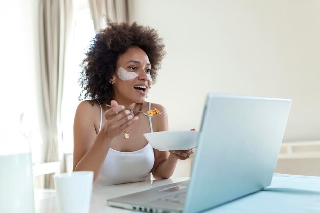 Attraktive Frau, die ihren Laptop-Computer benutzt, während sie zuhause frühstückt und zuhause lernt. Freiberufliches weibliches Modelmädchen, das morgens an ihrem Laptop arbeitet. Tasse Milch mit Cornflakes