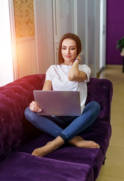 Attraktive Frau, die mit Laptop auf violettem Sofa sitzt. Überkreuzte Beine. Legerer Stil.