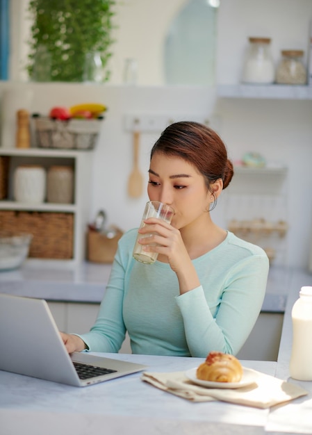 Attraktive Frau, die sich auf ihrem Laptop entspannt und posiert, während sie in ihrer Küche ein Glas Milch trinkt