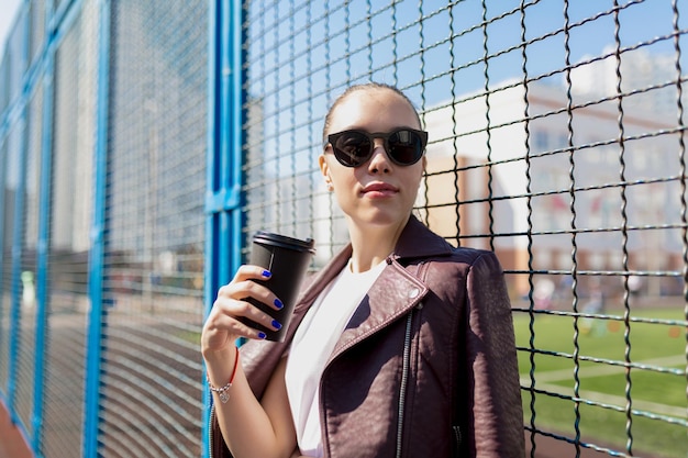 Attraktive Frau geht durch den städtischen Hintergrund Schöne Dame mit Sonnenbrille und burgunderfarbener Jacke lächelt und erkundet die Stadt am sonnigen Frühlingstag