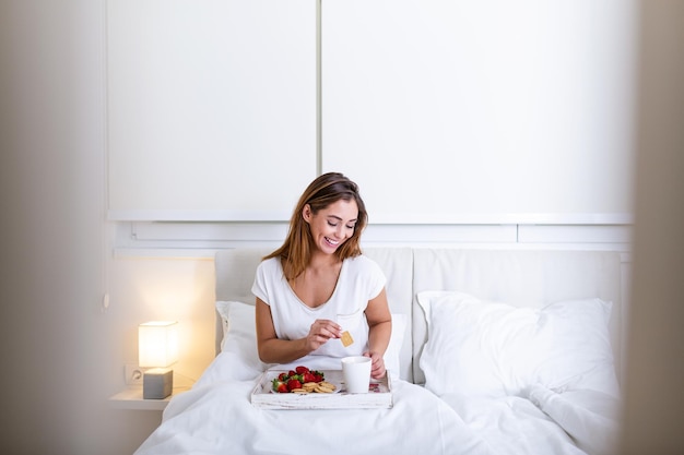 Attraktive Frau im Schlafanzug, die morgens mit dem Geruch von Kaffee und frischem Frühstück auf einem Holztablett im Bett aufwacht