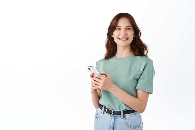 Attraktive Frau im T-Shirt mit Smartphone-Messanger, am Telefon chatten und in die Kamera lächeln, soziale Medien lesen, vor weißem Hintergrund stehen