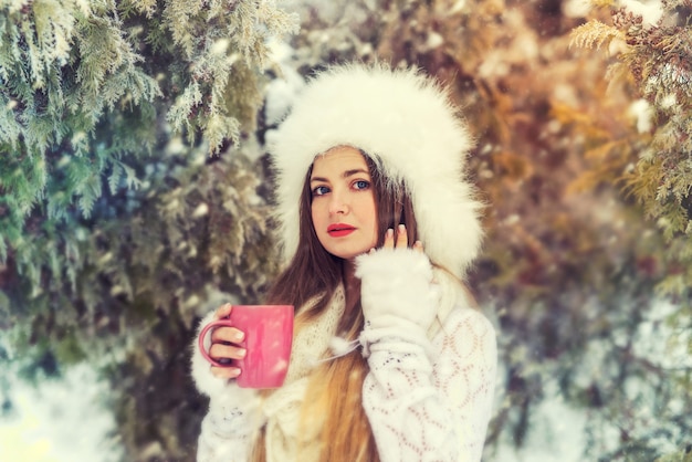 Attraktive Frau in gestricktem Pullover, Handschuhen und Mütze mit Tasse Kaffee im Winterwald