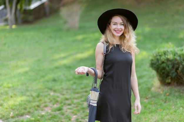 Attraktive Frau mit Filzhut und Kleid geht mit Regenschirm im Park spazieren