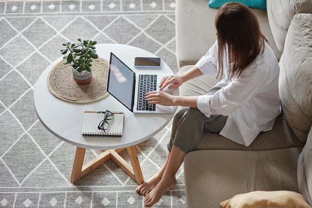 Attraktive Frau mit Laptop mit weißem Bildschirm, die zu Hause auf dem Sofa sitzt, Draufsicht