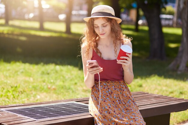 Attraktive Frau mit schickem Haar lädt Handy auf Bank mit Solarpanel auf, trinkt Kaffee und prüft E-Mails oder tippt Nachrichten