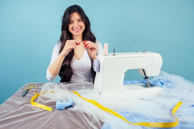 Attraktive Frau Schneider Hochzeitsschneiderin (Näherin) fädeln die Nadel an der Nähmaschine auf blauem Hintergrund im Studio. Das Konzept der Schaffung einer neuen Kollektion von Hochzeitskleidern