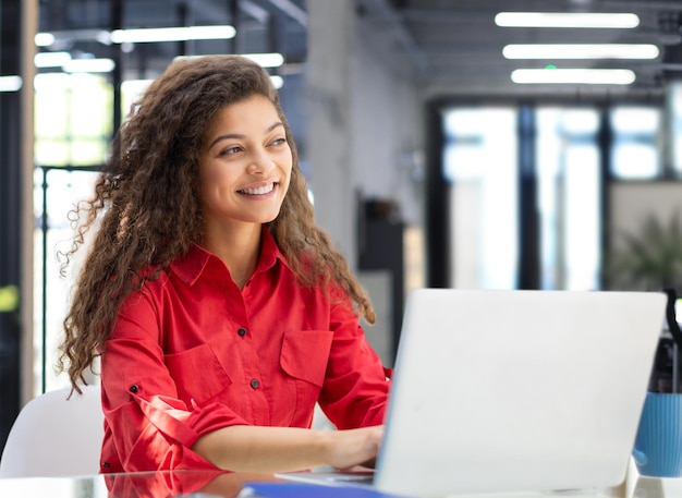 Attraktive, fröhliche Geschäftsfrau im roten Hemd, die im modernen Büro am Laptop arbeitet