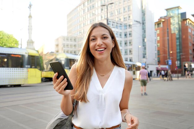 Attraktive Geschäftsfrau, die ein Smartphone hält, das auf einem städtischen Hintergrund in die Kamera blickt Taillenaufnahme