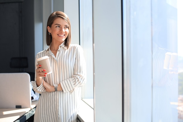 Attraktive Geschäftsfrau, die Kamera betrachtet und lächelt, während sie im Büro steht.