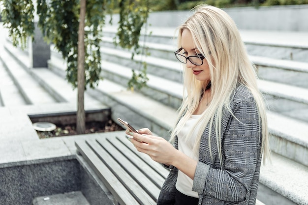 Attraktive Geschäftsfrau, die Smartphone in ihren Händen hält, während sie draußen auf einer Bank sitzt