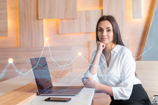 Attraktive Geschäftsfrau im weißen Hemd am Arbeitsplatz, die mit Laptop arbeitet, um die Handelsstrategie beim Corporate Finance Fund Forex-Chart-Hologramm über Bürohintergrund zu optimieren