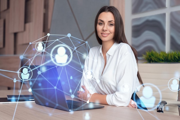 Attraktive Geschäftsfrau im weißen Hemd am Arbeitsplatz, die mit Laptop arbeitet, um neue Mitarbeiter für die internationale Unternehmensberatung HR-Social-Media-Hologrammsymbole über Bürohintergrund einzustellen