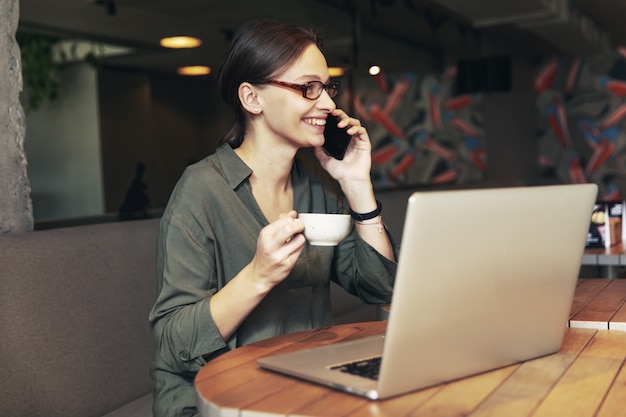 Attraktive Geschäftsfrau in schwarzer Jacke und stilvoller Brille, die im Café sitzt, am Telefon spricht und Laptop betrachtet.