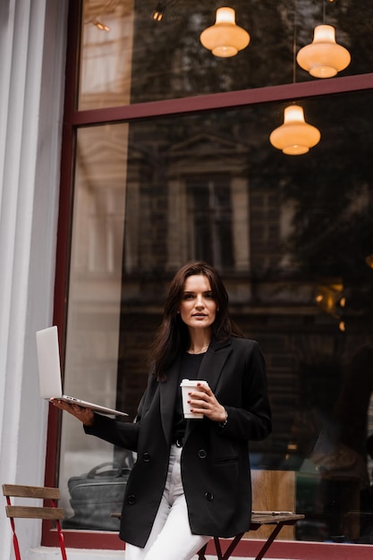 Attraktive Geschäftsfrau mit Laptop steht in der Nähe des Cafés Selbstbewusstes Modellmädchen steht und posiert mit Laptop und einer Tasse Kaffee außerhalb des Cafés