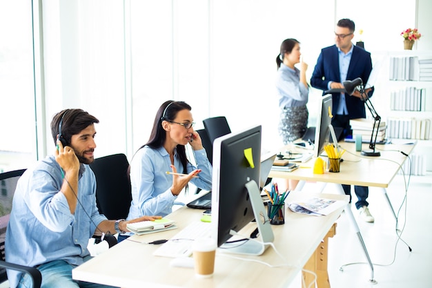 Attraktive Geschäftsleute in Headsets lächeln, während sie in einem modernen Büro mit dem Computer arbeiten