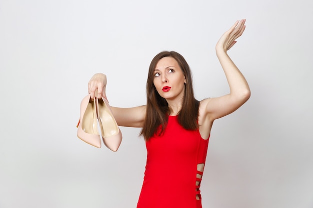 Attraktive Glamour-kaukasische modische junge nachdenkliche, verwirrte braunhaarige Frau im roten Kleid, die beige Schuhe mit roter Einkaufssohle einzeln auf weißem Hintergrund bekommt. Kopieren Sie Platz für Werbung.