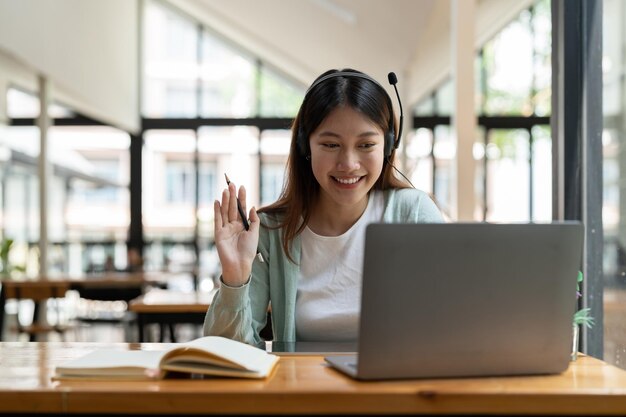 Attraktive, glückliche junge Studentin, die zu Hause online mit Laptop-Kopfhörern studiert und Video-Chat winkt Remote Work Distance Education Videokonferenz oder virtuelle Veranstaltung zur Quarantäne