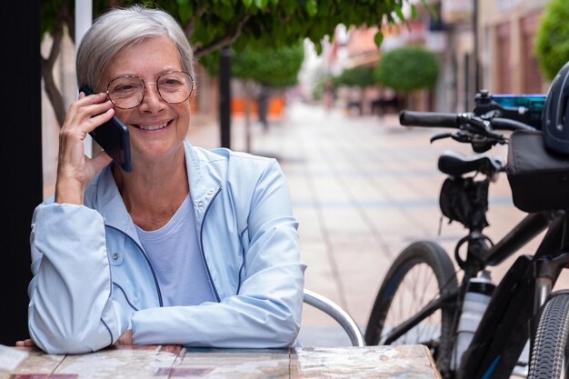 Attraktive grauhaarige Seniorin, die ein Smartphone benutzt, während sie an einem Tisch im Café im Freien sitzt Ältere Frau, die in der Nähe ihres Elektrofahrrads sitzt und Technik und Soziales genießt