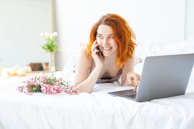 Attraktive Ingwerfrau mittleren Alters im Pyjama, die im Bett mit Telefon und Laptop liegt.