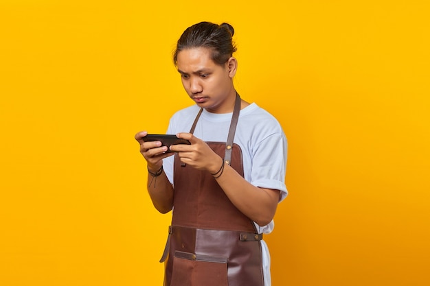 Attraktive junge asiatische Barista mit Handy-Spiel auf gelbem Hintergrund