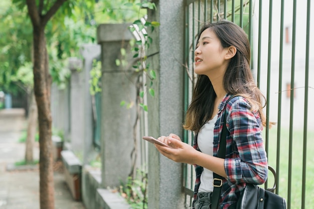 Attraktive junge asiatische Frau, die im Schatten unter Bäumen steht und sich an einen Metallzaun lehnt. süßes mädchen, das handy überprüft und wegschaut. schöne Frau mit Handy und entspannen Sie sich im Freien.