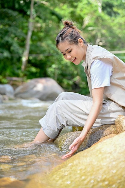 Attraktive junge asiatische Frau, die sich entspannt in der Nähe des Flusses sitzt und das Süßwasser berührt