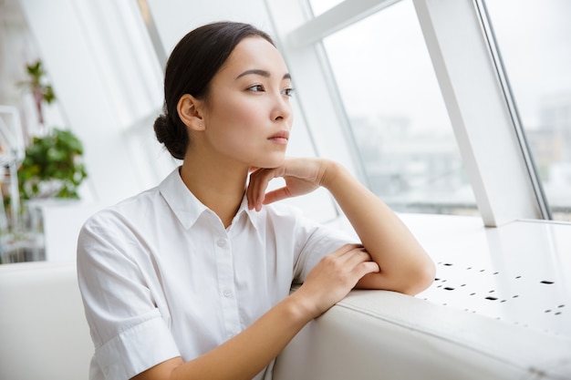 Attraktive junge asiatische Geschäftsfrau, die sich auf eine Couch stützt, während sie im Café drinnen sitzt