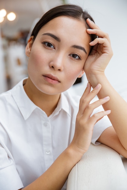 Attraktive junge asiatische Geschäftsfrau, die sich auf eine Couch stützt, während sie im Café drinnen sitzt