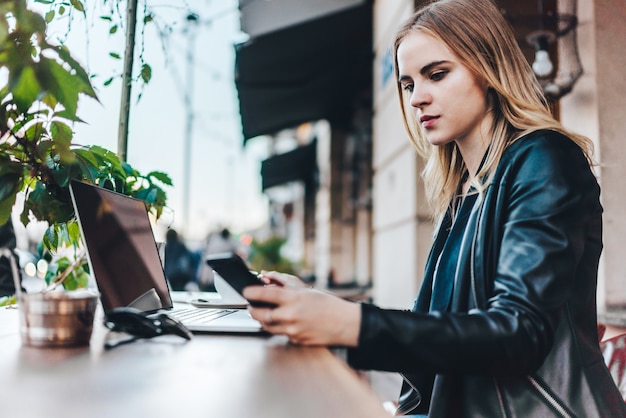 Attraktive junge blonde Geschäftsfrau gekleidet in Lederjacke, die ihre Mittagspause auf der Straßencaféterrasse verbringt und an ihrem Laptop arbeitet