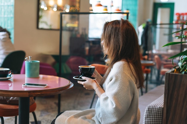 Attraktive junge brünette lächelnde Frau in weißem Freizeitkleid mit einer Tasse Kaffee in den Händen im Café