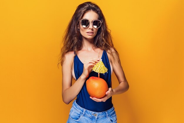 Attraktive junge Dame im Sommeroutfit, die frische saftige Zitrusfrüchte mit Strohhalm und Regenschirm hält und Kamera beim Stehen auf Gelb betrachtet