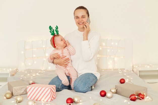 Attraktive junge erwachsene glückliche Frau mit positivem Ausdruck, die auf dem Bett mit Baby in den Armen sitzt und Telefon spricht und jemandem zu Weihnachten und guten Rutsch ins Neue Jahr gratuliert.