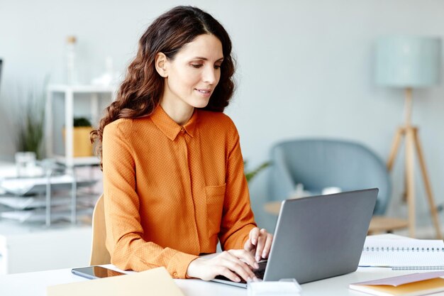 Attraktive junge erwachsene kaukasische Frau, die eine stilvolle orangefarbene Bluse trägt, sitzt an einem Schreibtisch und tippt auf einem Laptop