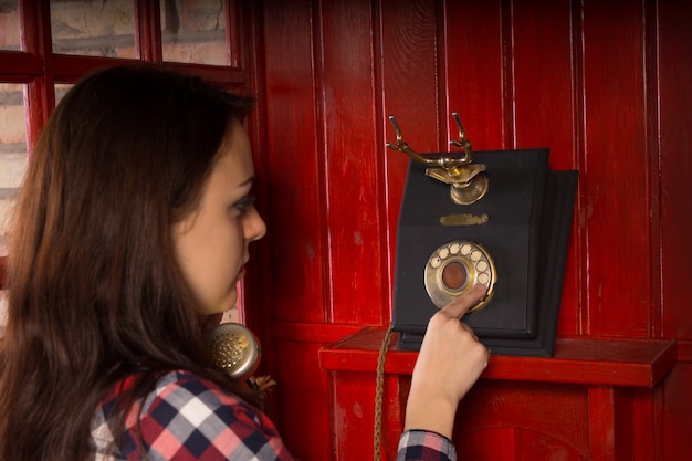 Attraktive junge Frau, die an einem alten Vintage-Wähltelefon in einer roten Telefonzelle aus Holz telefoniert