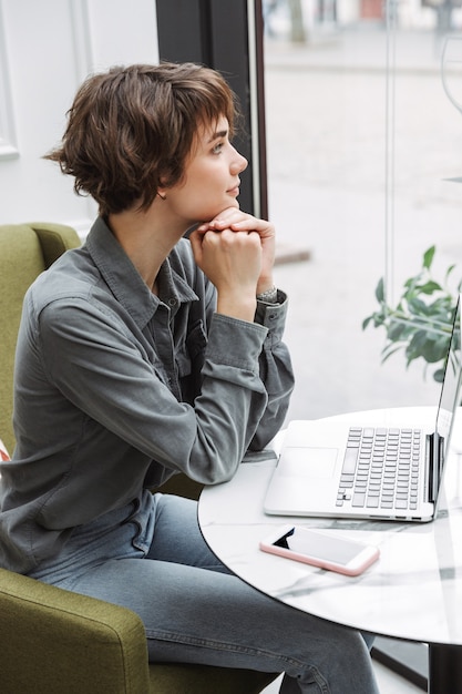 Attraktive junge Frau, die drinnen am Café-Tisch sitzt, am Laptop arbeitet und Dokumente analysiert