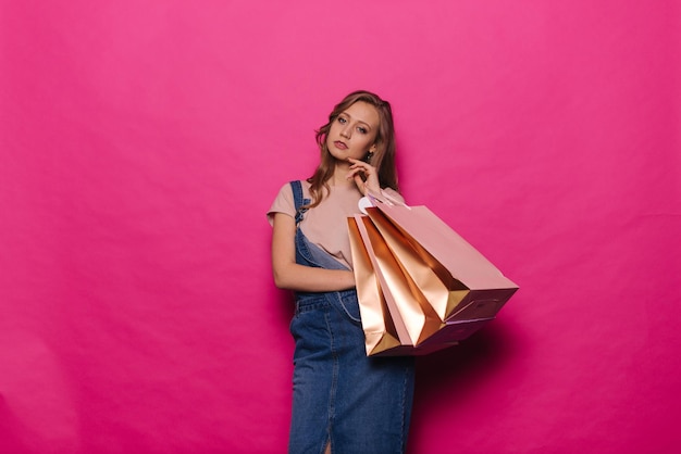 Attraktive junge Frau, die goldene Papiertüten hält, nachdem sie im Einkaufszentrum auf isoliertem rosa Hintergrund eingekauft hat. Fröhliches Mädchen in Denim-Kleidung posiert mit Paketen im Saisonverkauf im professionellen Studio.