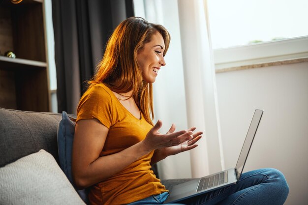 Attraktive junge Frau, die im Schneidersitz auf dem Sofa sitzt und mit ihrem Laptop einen Video-Chat mit jemandem zu Hause macht.