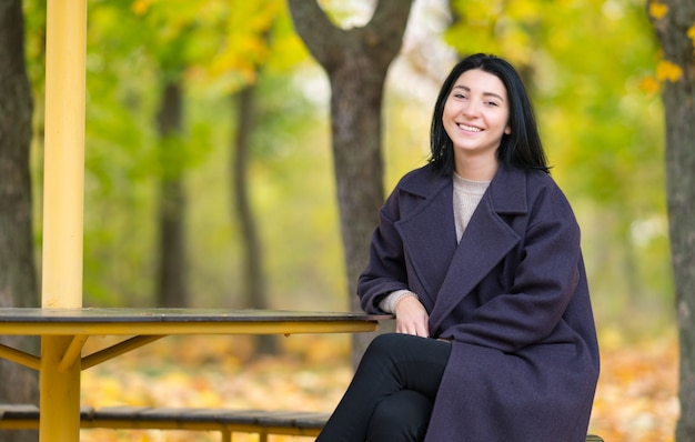 Attraktive junge Frau, die in einem Park im Herbst sitzt, der auf einer Bank sitzt, die Kamera mit einem warmen freundlichen Lächeln betrachtet