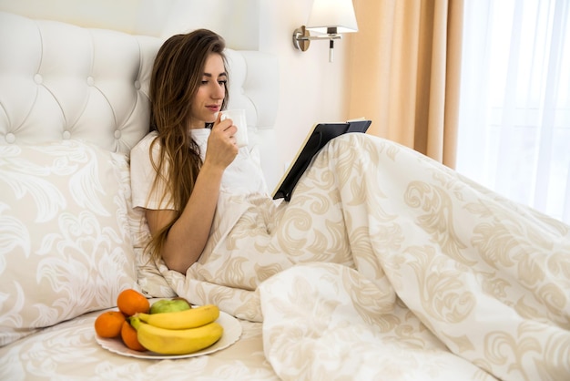 Attraktive junge Frau, die morgens Kaffee mit Obst trinkt, liest ihr Notizbuch im Bett. Wochenendkonzept