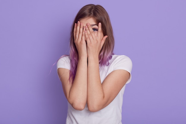 Attraktive junge Frau, die weißes lässiges T-Shirt trägt, das ihr Gesicht mit Handflächen bedeckt und durch Finger schaut, für jemanden spioniert, lokalisiert über lila Wand.