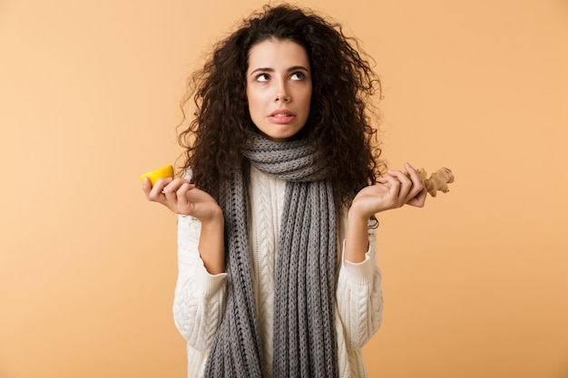 Attraktive junge Frau, die Winterschal trägt, der Ingwer und Zitrone zeigt, während lokalisiert über weißer Wand steht
