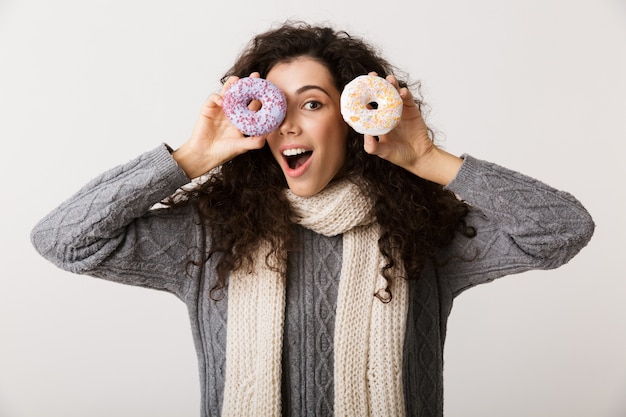 Attraktive junge Frau, die Winterschal trägt, der süße Donuts zeigt, während lokalisiert über weißer Wand steht