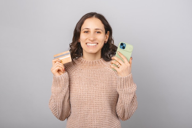 Attraktive junge Frau hält ein Telefon und eine Kreditkarte auf grauem Hintergrund