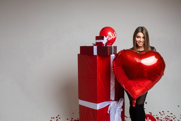 Attraktive junge Frau hält einen Luftballon in ihren Händen und steht in der Nähe von Geschenken, Bild auf Weiß