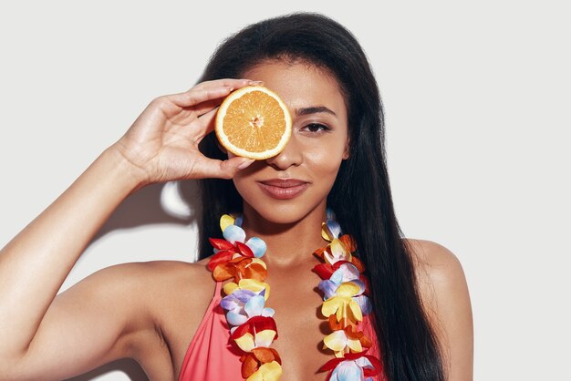 Attraktive junge Frau im Bikini, die das Auge mit einer Orangenscheibe bedeckt und lächelt, während sie vor grauem Hintergrund steht
