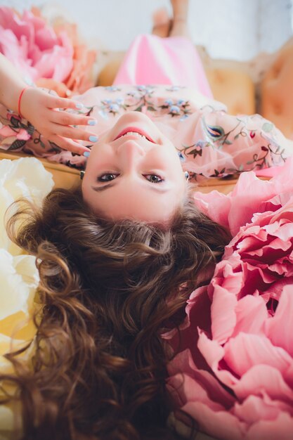 Attraktive junge Frau im eleganten Kleid, das im Innenraum mit Blumen verziert sitzt. Schönes frisches lächelndes Mädchen, das mit Blumen aufwirft