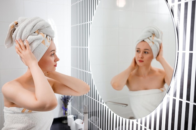Attraktive junge Frau im Handtuch schaut nach dem Duschen auf den Spiegel, der im Badezimmer steht Schönheits- und Hautpflegekonzept