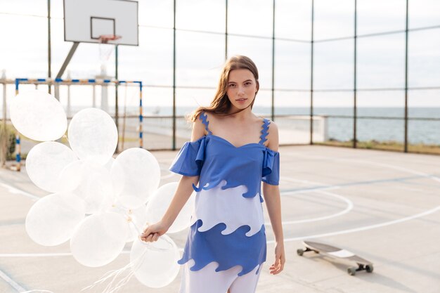 Attraktive junge Frau im stilvollen Kleid, das Luftballons auf Sportplatz im Freien steht und hält