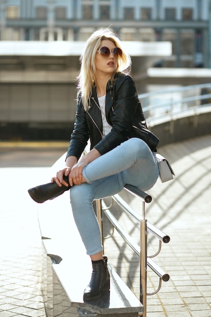 Attraktive junge Frau in Jeans und Jacke, die am Sommertag auf der Straßenbrüstung sitzt